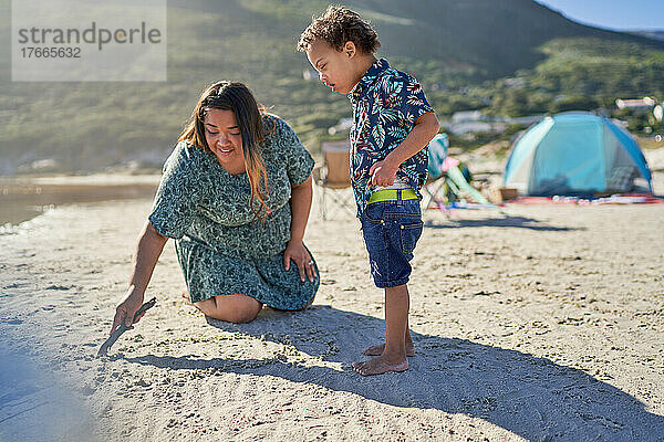 Mutter und Sohn mit Down-Syndrom zeichnen mit Stock im Sand am Strand