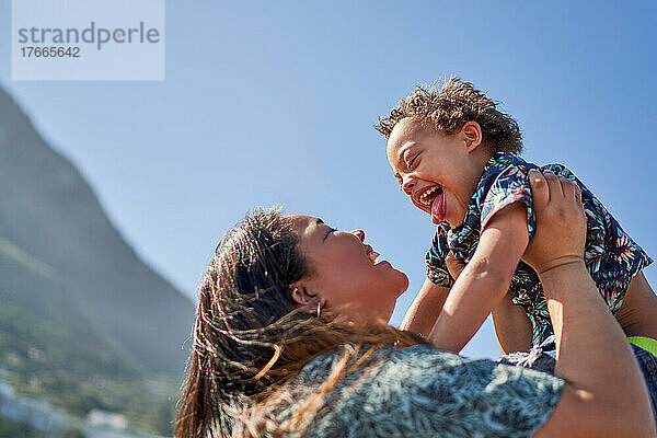 Glückliche Mutter hebt ihren Sohn mit Down-Syndrom unter den sonnigen blauen Himmel