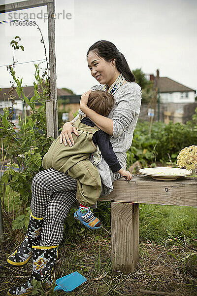 Glückliche Mutter hält müden Kleinkindsohn auf Bank im Garten