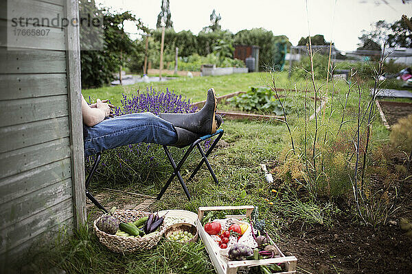 Mann entspannt sich mit erhobenen Füßen im Garten neben geerntetem Gemüse