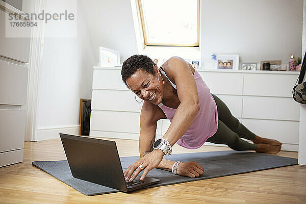 Glückliche reife Frau  die am Laptop auf einer Yogamatte trainiert