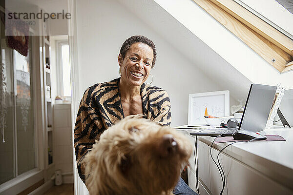 Porträt einer glücklichen Frau mit Hund am Schreibtisch im Heimbüro