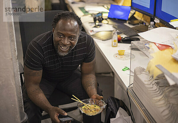 Lächelnder Geschäftsmann isst Mittagessen zum Mitnehmen am Schreibtisch