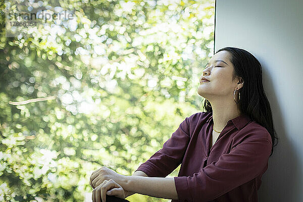 Ruhige junge Frau entspannt sich am Fenster mit Blick auf den grünen Baum
