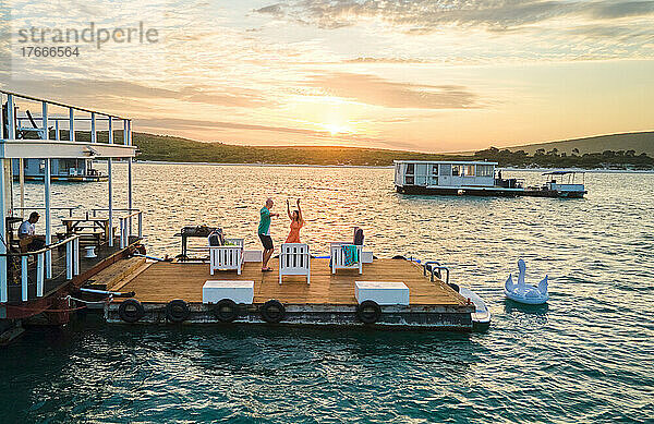 Unbeschwertes Paar tanzt bei Sonnenuntergang auf der Terrasse des Hausboots am See