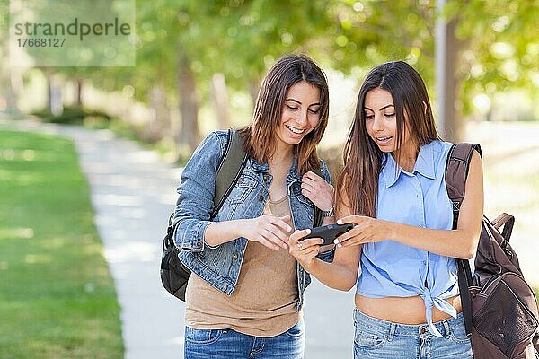 Zwei schöne junge ethnische Zwillingsschwestern mit Rucksäcken  die draußen ein Smartphone benutzen