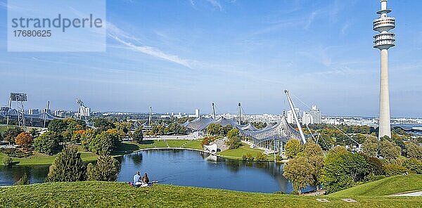 Zwei Personen sitzen im Park mit Olympiasee und Olympiaturm  Olympiapark München  Bayern  Deutschland  Europa