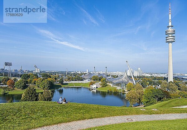 Zwei Personen sitzen im Park mit Olympiasee und Olympiaturm  Olympiapark München  Bayern  Deutschland  Europa