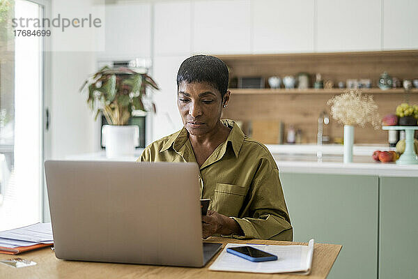 Reife Frau hält Smartphone in der Hand und benutzt Laptop