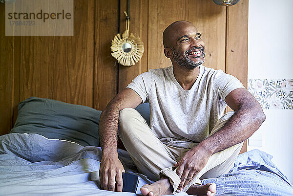 Lächelnder Mann hält Smartphone in der Hand  während er im Schlafzimmer sitzt