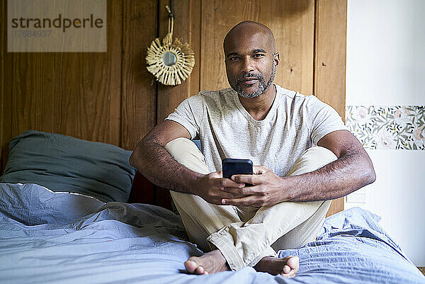 Lächelnder Mann hält Smartphone in der Hand  während er im Schlafzimmer sitzt