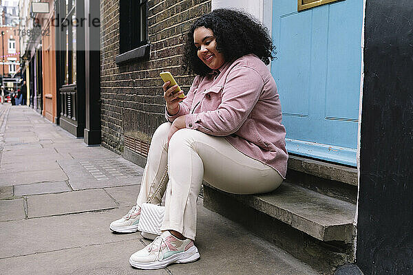 Lächelnde Frau  die Textnachrichten über ihr Smartphone sendet und auf einer Stufe sitzt