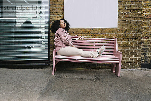 Junge Frau mit geschlossenen Augen sitzt auf rosa Bank