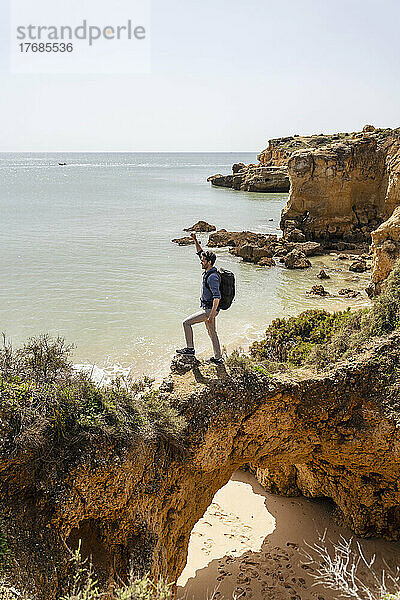 Sorgloser Mann mit erhobener Hand steht auf einer Felsbrücke vor dem Meer