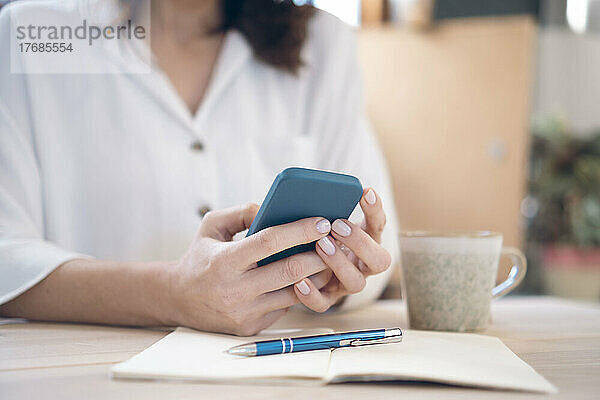 Frau mit Smartphone und Tagebuch sitzt zu Hause am Tisch