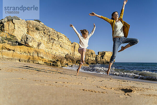 Glückliche Frau mit Tochter springt an einem sonnigen Tag am Strand