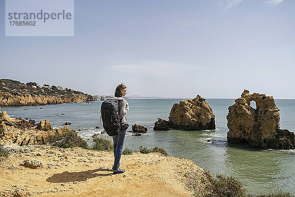 Frau mit Rucksack steht an einem sonnigen Tag vor dem Meer und blickt auf die Aussicht