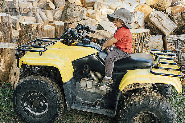 Junge mit Hut sitzt auf einem Quad vor Holzstämmen