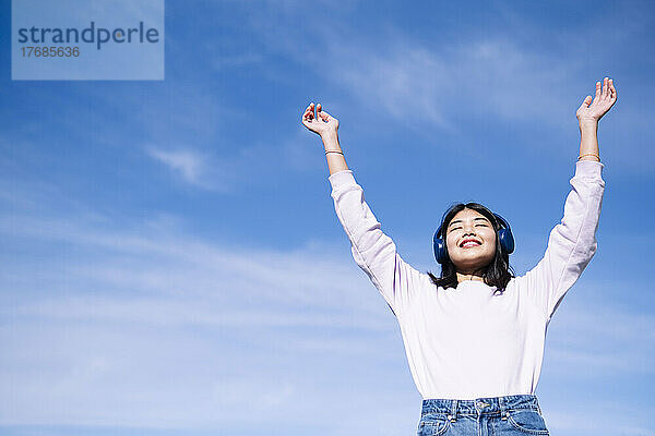 Glückliche Frau hört Musik mit erhobenen Händen vor dem Himmel