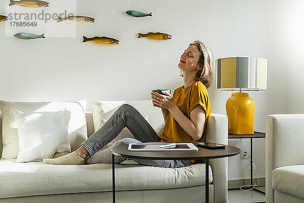 Lächelnde Frau mit geschlossenen Augen und einer Kaffeetasse  die zu Hause auf dem Sofa sitzt