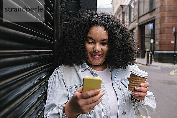 Lächelnde junge Frau hält eine Einweg-Kaffeetasse in der Hand und nutzt ihr Smartphone vor dem Auslöser