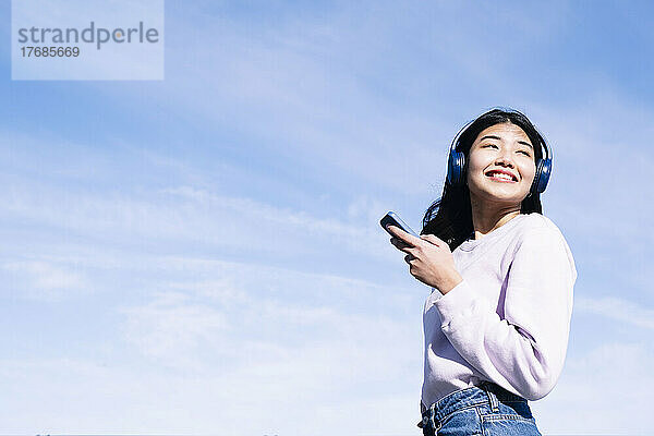 Lächelnde Frau mit Kopfhörern und Smartphone an einem sonnigen Tag