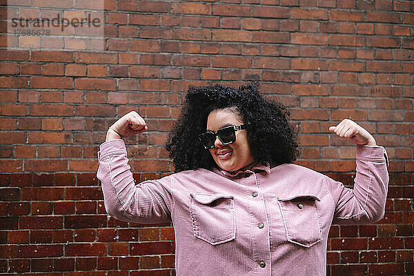 Smiling woman wearing sunglasses flexing muscles in front of wall