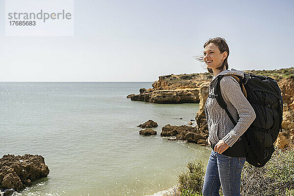 Glückliche Frau mit Rucksack  die an einem sonnigen Tag vor dem Meer steht