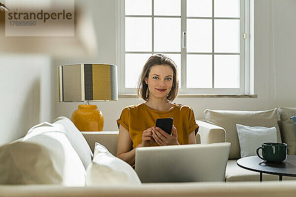 Lächelnder Freiberufler mit Smartphone und Laptop sitzt auf dem Sofa im Wohnzimmer