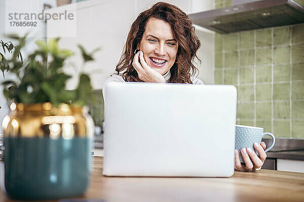 Glücklicher Freiberufler  der mit der Hand am Kinn sitzt und zu Hause einen Laptop benutzt