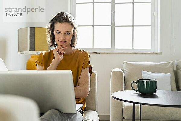 Lächelnder Freiberufler mit der Hand am Kinn und Blick auf den Laptop im Wohnzimmer