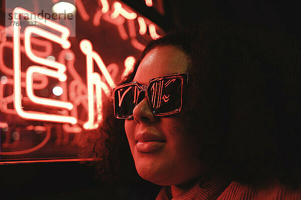 Junge Frau mit Sonnenbrille an beleuchteter Wand in der Nacht