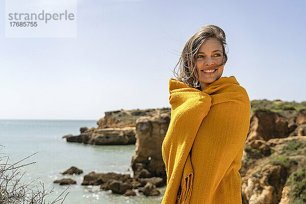 Glückliche Frau  in eine Decke gehüllt  steht vor dem Meer