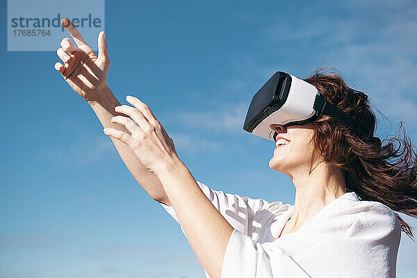 Glückliche Frau mit Virtual-Reality-Simulator  die an einem sonnigen Tag vor dem Himmel steht