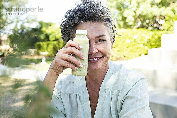 Lächelnde Frau bedeckt ihr Gesicht mit einer Flasche Smoothie und sitzt im Garten