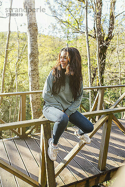 Lächelnde junge Frau sitzt auf dem Holzgeländer der Brücke