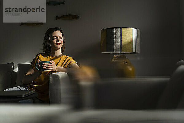 Lächelnde Frau mit geschlossenen Augen und Kaffeetasse im Wohnzimmer sitzend