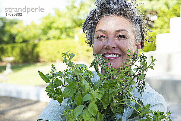 Lächelnde reife Frau mit Pflanze sitzt an einem sonnigen Tag im Garten