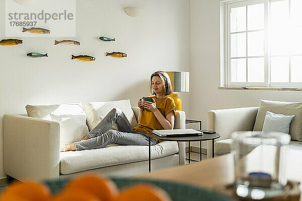 Frau mit Kaffeetasse sitzt auf Sofa im Wohnzimmer