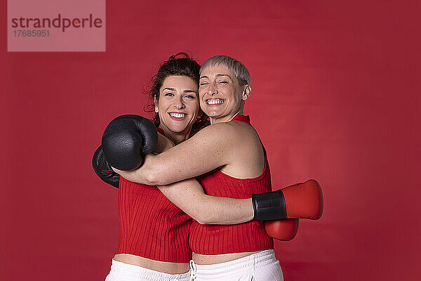 Lächelnde Freunde mit Boxhandschuhen  die sich vor rotem Hintergrund umarmen