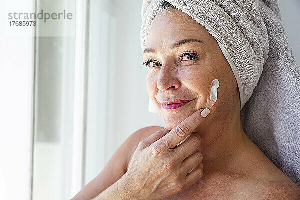 Lächelnde reife Frau trägt zu Hause Feuchtigkeitscreme auf ihr Gesicht auf