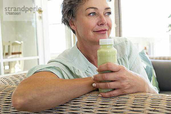 Lächelnde reife Frau mit einer Flasche Smoothie sitzt zu Hause auf dem Sofa