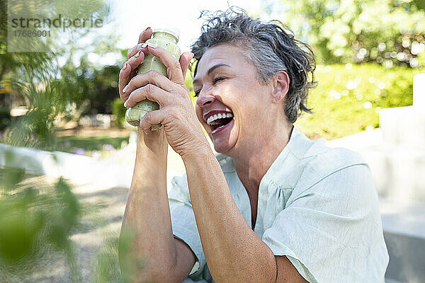 Fröhliche Frau mit einer Flasche Smoothie sitzt im Garten