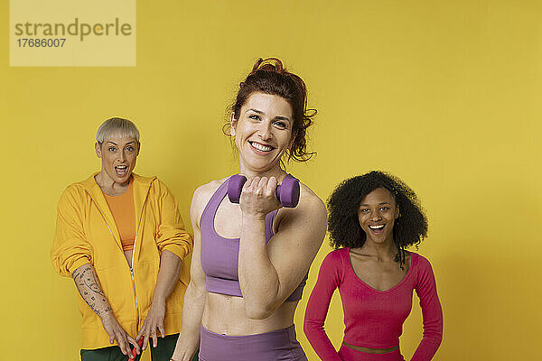 Lächelnde Frau hält Hantel und steht mit Freunden vor gelbem Hintergrund