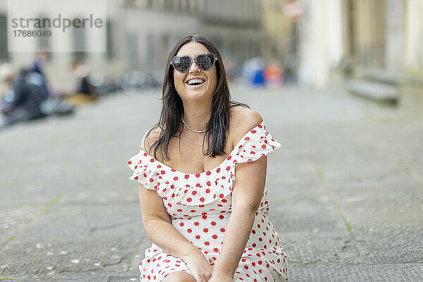 Glückliche Frau mit herzförmiger Sonnenbrille sitzt auf Fußweg