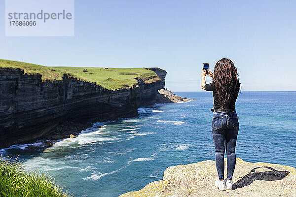 Junge Frau fotografiert das Meer mit ihrem Mobiltelefon  das auf einem Felsen steht