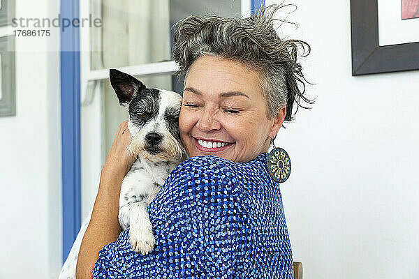 Glückliche reife Frau mit kurzen Haaren umarmt Hund zu Hause