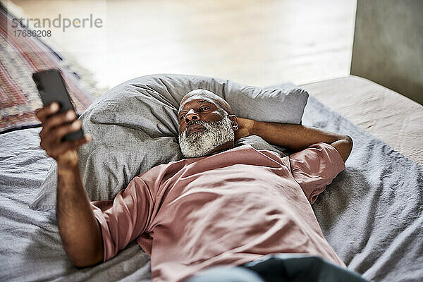 Glatzköpfiger Mann mit der Hand hinter dem Kopf und Smartphone  der zu Hause im Bett liegt
