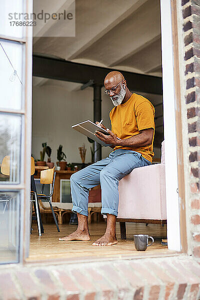 Glatzköpfiger Mann benutzt Tablet-PC und sitzt zu Hause auf dem Sofa