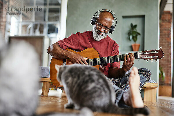 Reifer Mann mit Kopfhörern  der zu Hause Gitarre für Katzen spielt
